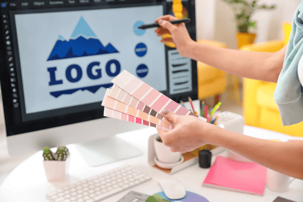 A lady at a desk,  holding pink colour swatches in one hand and a digital pen in the other, designing a logo for printing, on her PC screen.