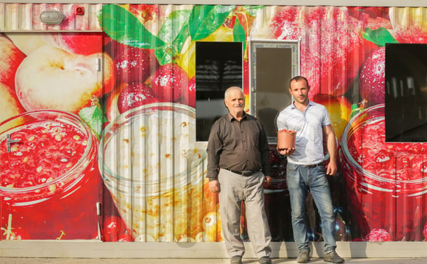 Two men standing in front of a large digital printed wall featuring colourful fruit juice jars and fresh fruit graphics on a container building exterior.