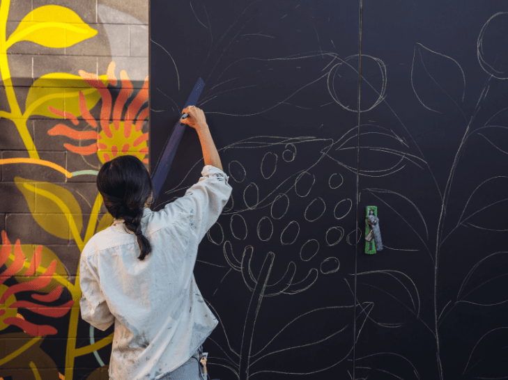 A Female artist creating a custom wall mural by drawing an outline on a blank wall, and filling it in with a specific colour pallet.