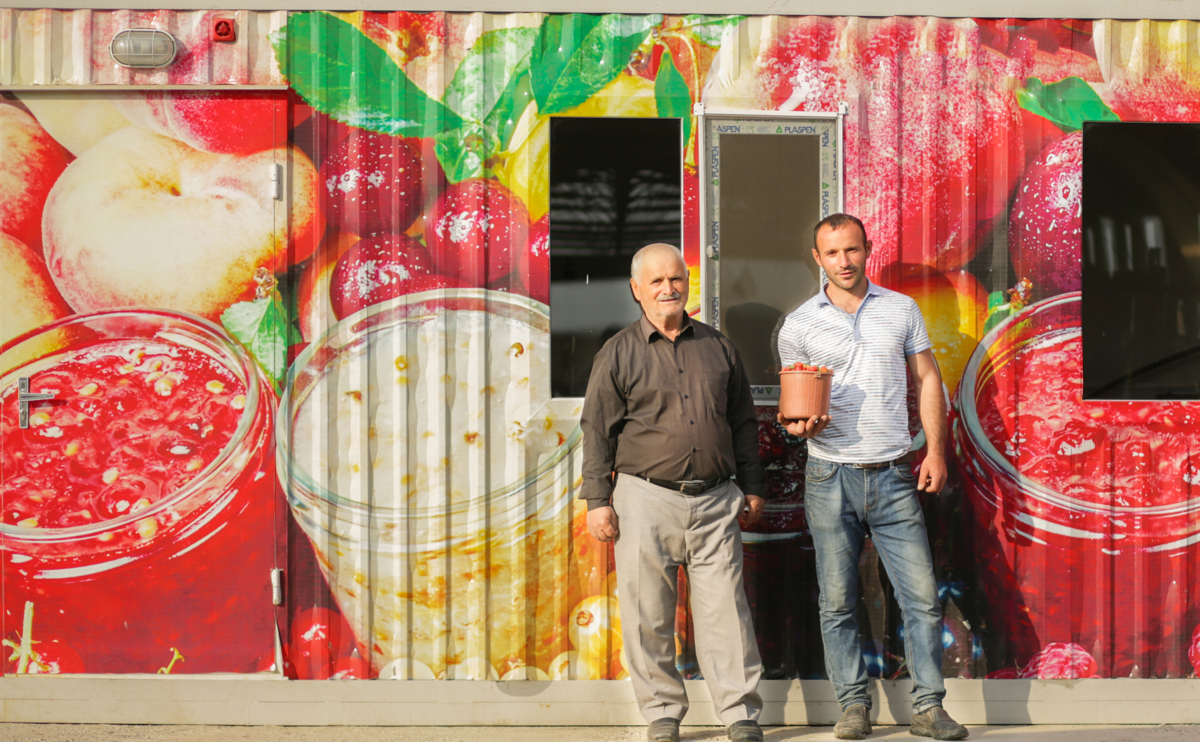 Two men standing in front of a large digital printed wall featuring colourful fruit juice jars and fresh fruit graphics on a container building exterior.