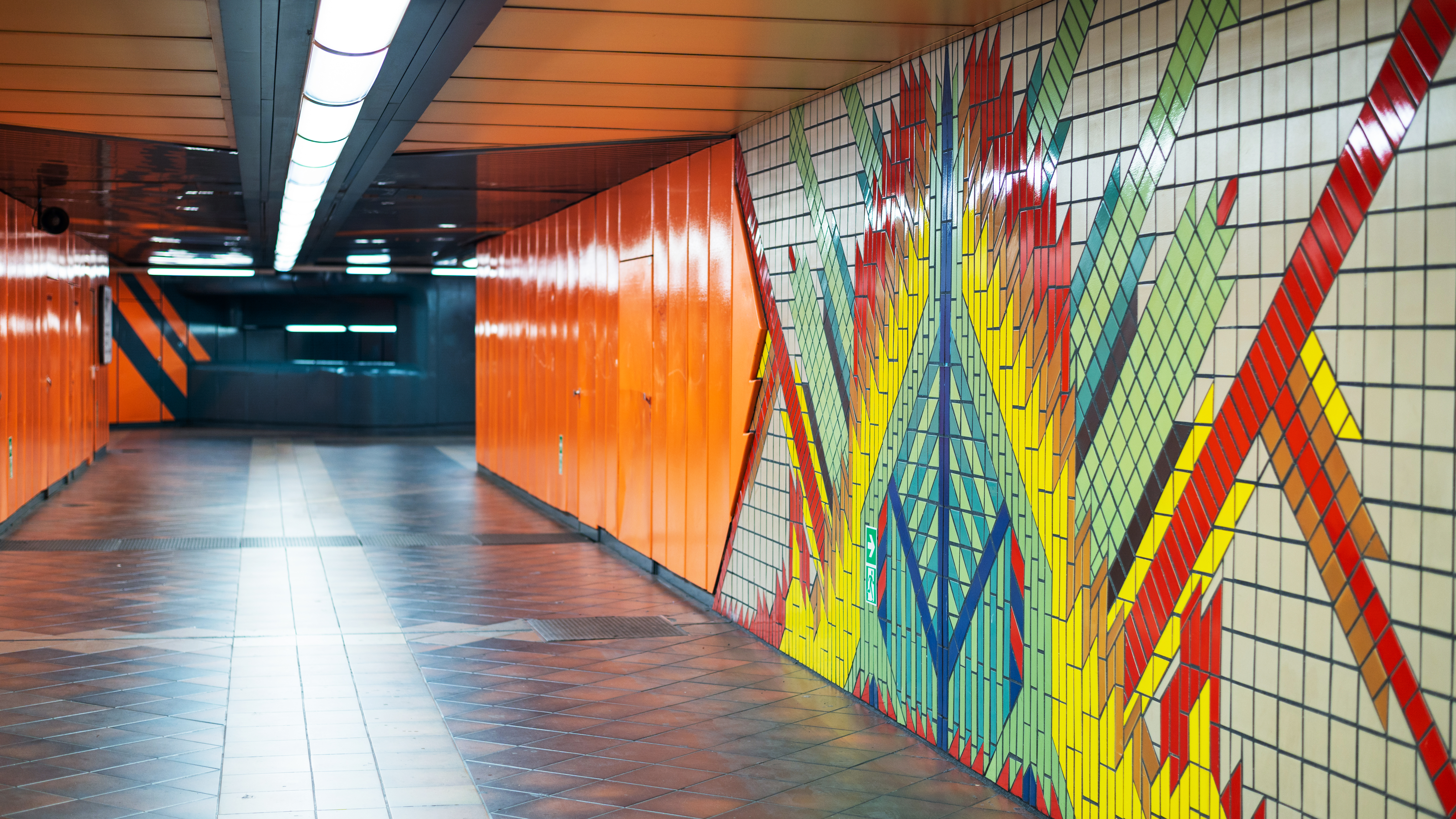 Colourful tiled wall graphics mural in an underground station corridor with patterned flooring and modern lighting.